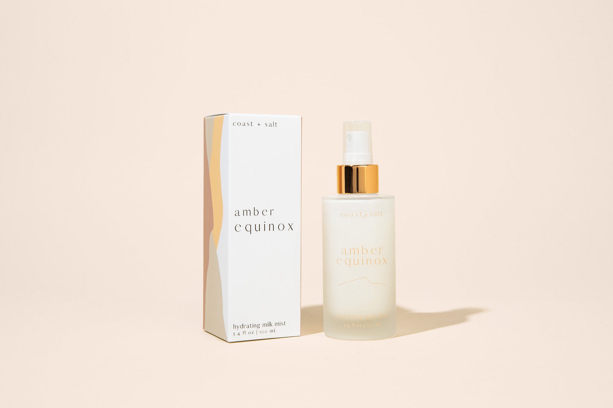 A frosted glass spray bottle of spray lotion labeled amber equinox next to its white and gold packaging box, set against a solid pale peach background.