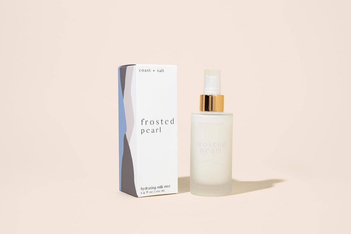 A bottle of sprayable lotion labeled "Frosted Pearl" next to its product packaging on a beige background