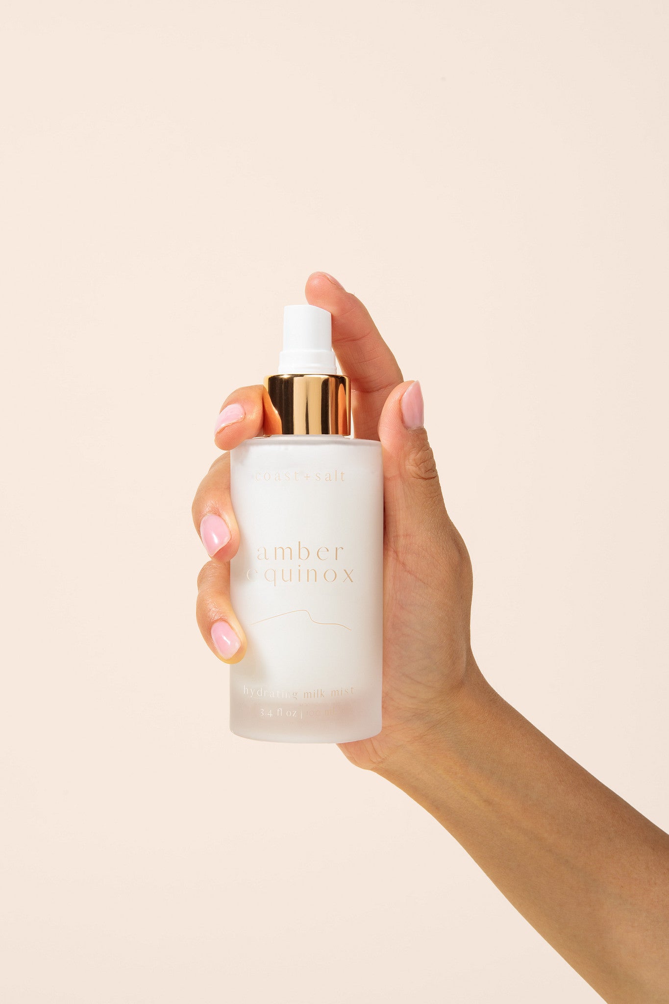A hand with pink manicured nails holds a frosted glass spray bottle with a metallic gold collar and white spray top. The bottle has gold lettering that reads 'amber equinox'.
