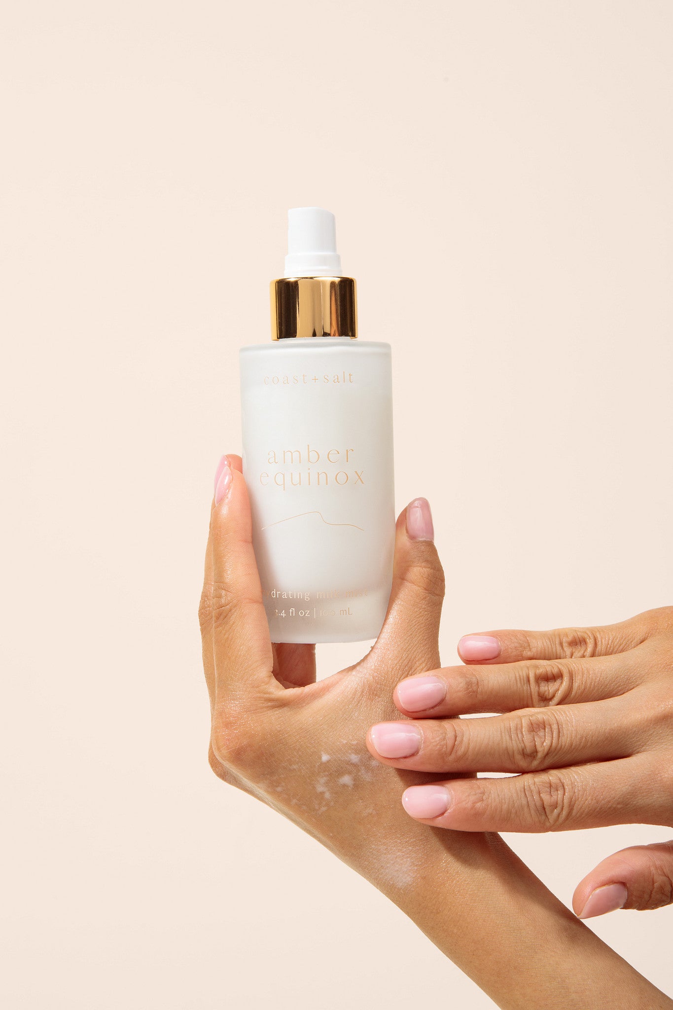 A frosted glass pump bottle labeled 'amber equinox' with a white cap and gold collar, held by one hand while another hand rubs in a lotion product onto the first hand against a soft beige background.