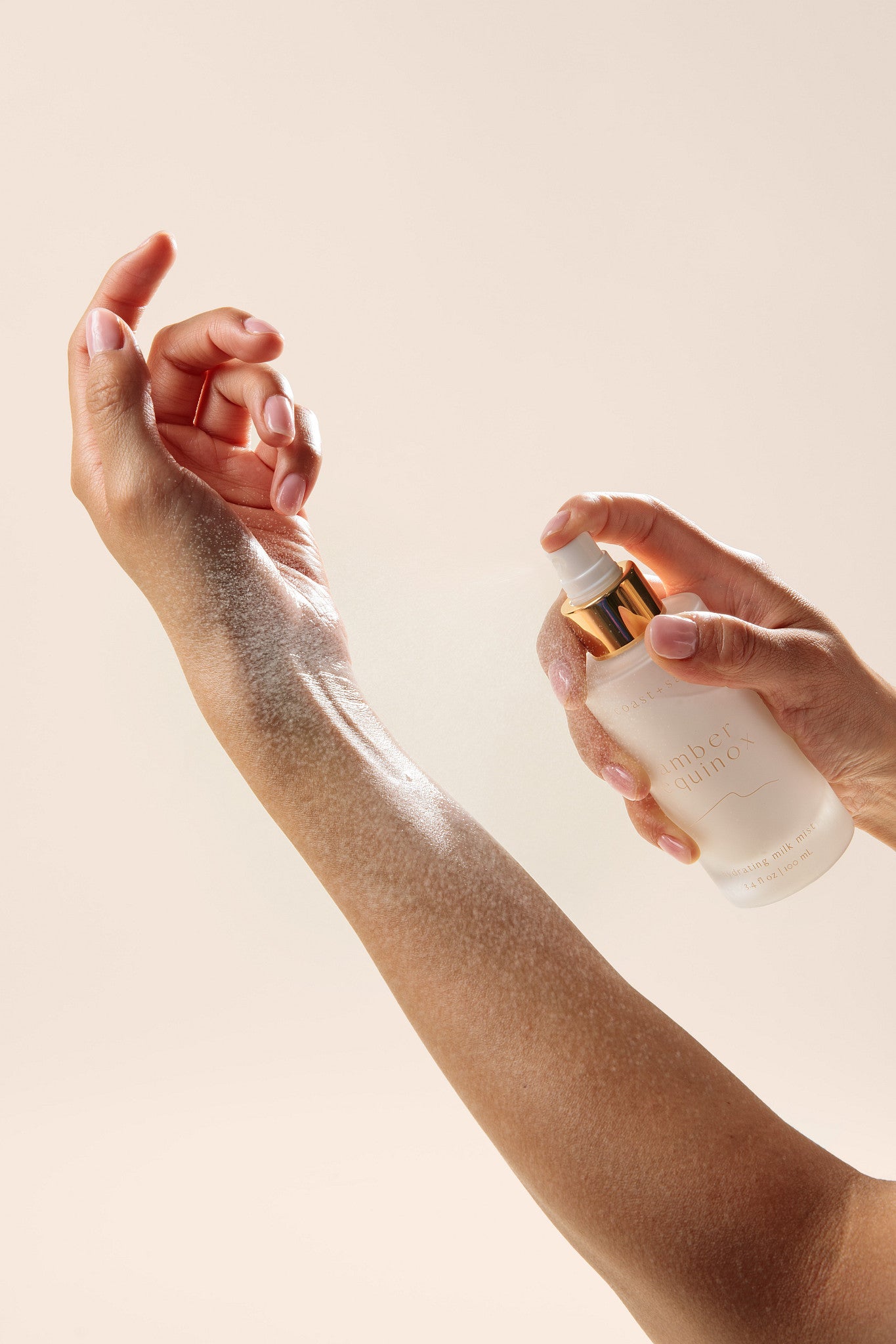 A close-up of a person's arm and hand spraying a milky skincare product onto her arm, the bottle is frosted with a gold accent collar, against a solid light beige background.