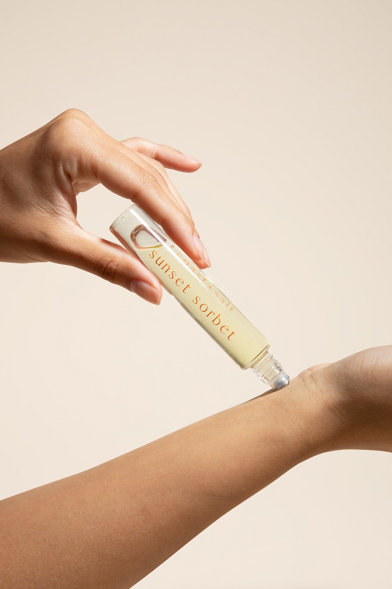 Hand applying a clear bottle labeled 'Sunset Sorbet' above an arm on a beige background