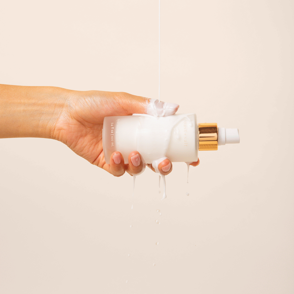 Hand holding a frosted Amber Equinox Hydrating Milk Mist bottle as milky liquid pours over the bottle.