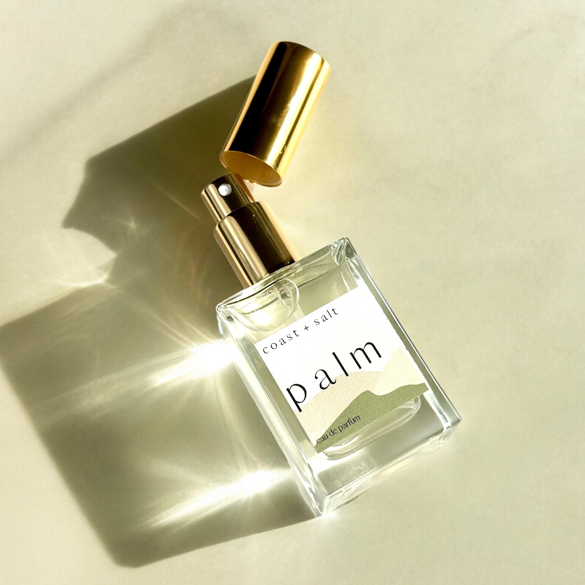 A bottle of Palm Perfume laying flat on a beige concrete surface. The gold cap is off and laying diagonally above the gold atomizer sprayer.