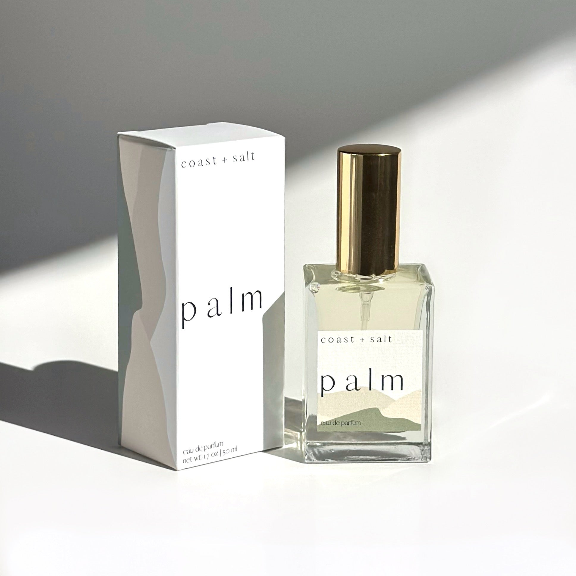 A bottle of Palm Perfume next to the Palm Perfume Retail Box. The box and the perfume bottle are lit in soft daylight, and sitting on a white surface with a diagonal  shadow in the background.