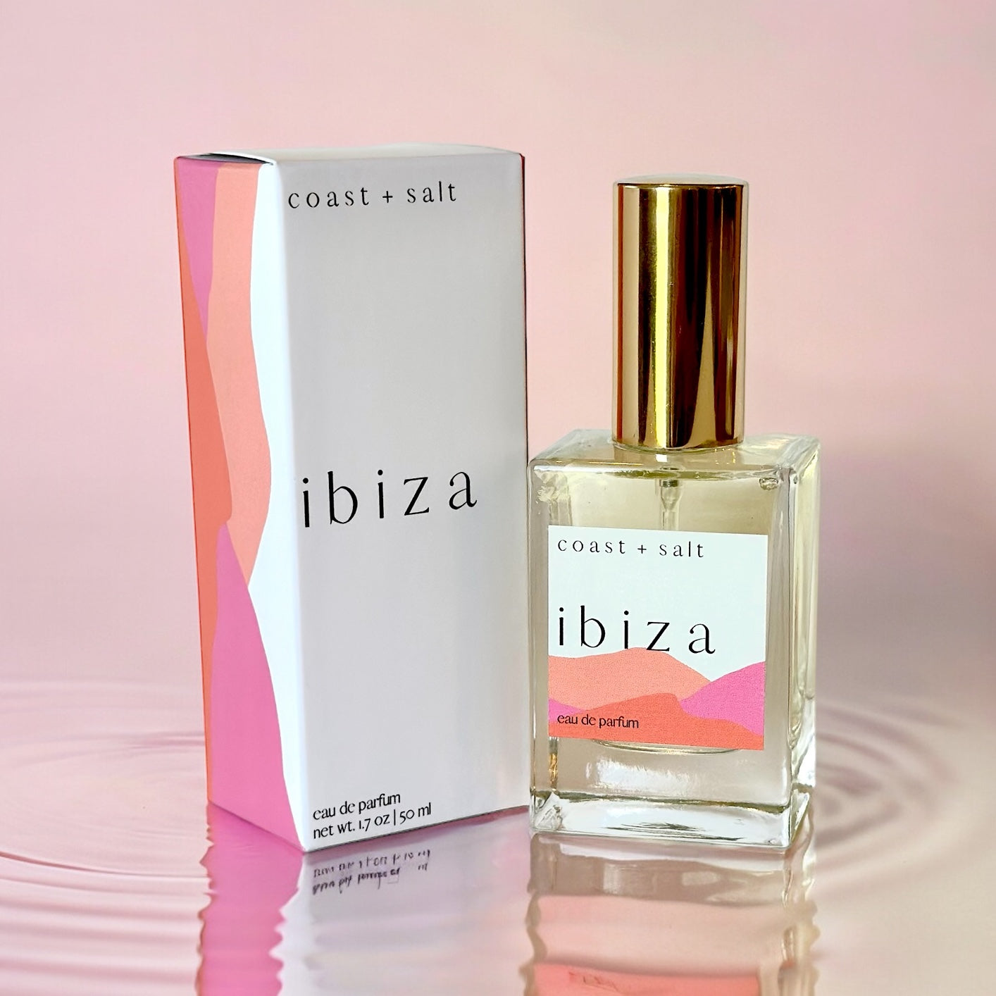 A bottle of Ibiza Perfume next to the Ibiza Perfume Retail Box. The box and the perfume bottle are sitting on top of rippling water in a light pink hue.