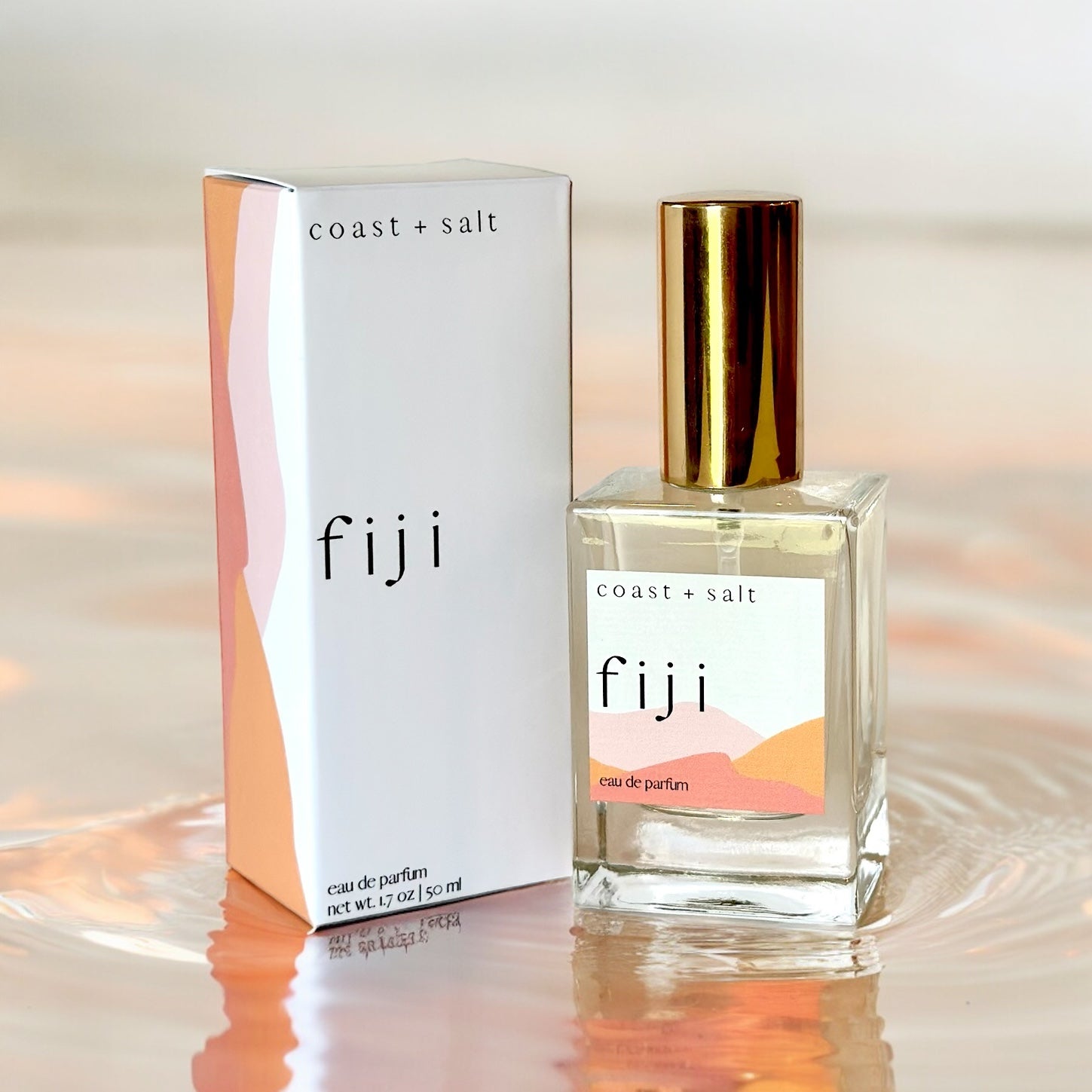 A bottle of Fiji Perfume next to the Fiji Perfume Retail Box. The box and the perfume bottle are sitting on top of rippling water in a light pink hue.