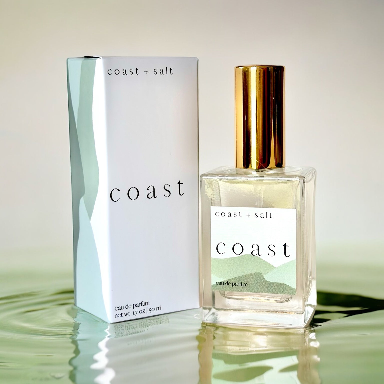 A bottle Of Coast Perfume next to the Coast Perfume Retail Box. The box and the perfume bottle are sitting on top of rippling water in a green hue.