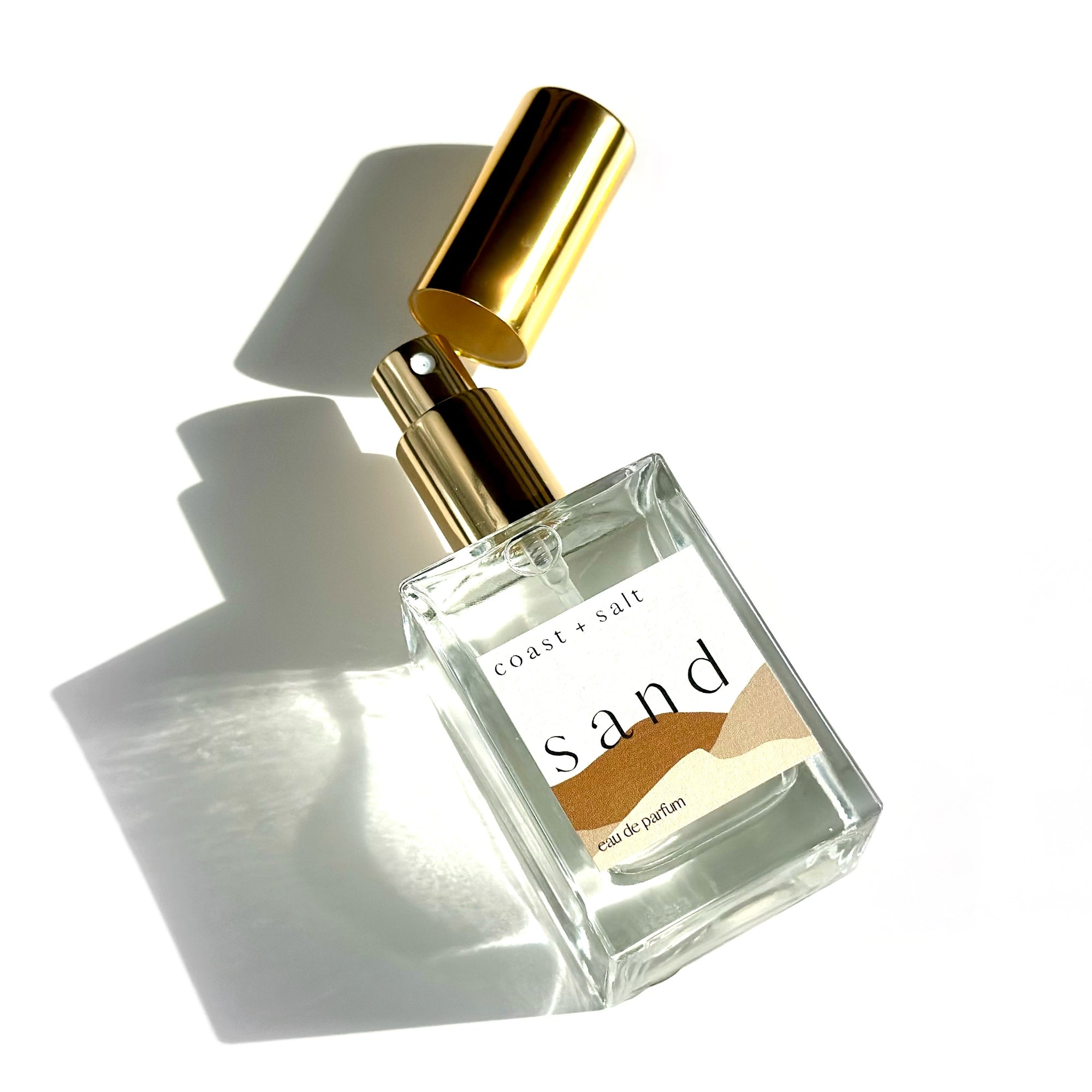 A bottle of Sand Perfume laying flat on a white surface. The gold cap is off and laying diagonally above the gold atomizer sprayer.