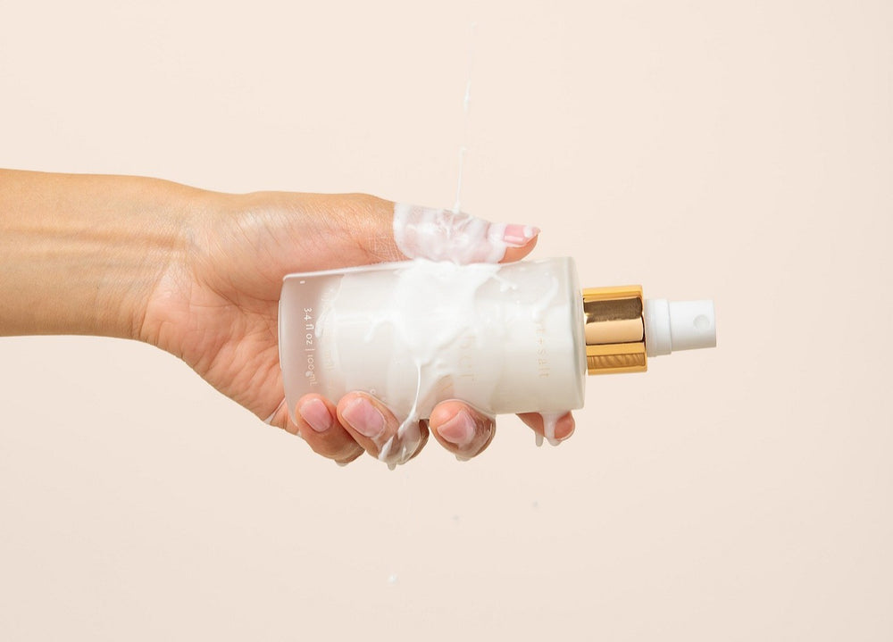 A hand holding a frosted glass bottle with a white pump and gold collar, with white cream dripping onto the bottle, against a solid pale beige background.